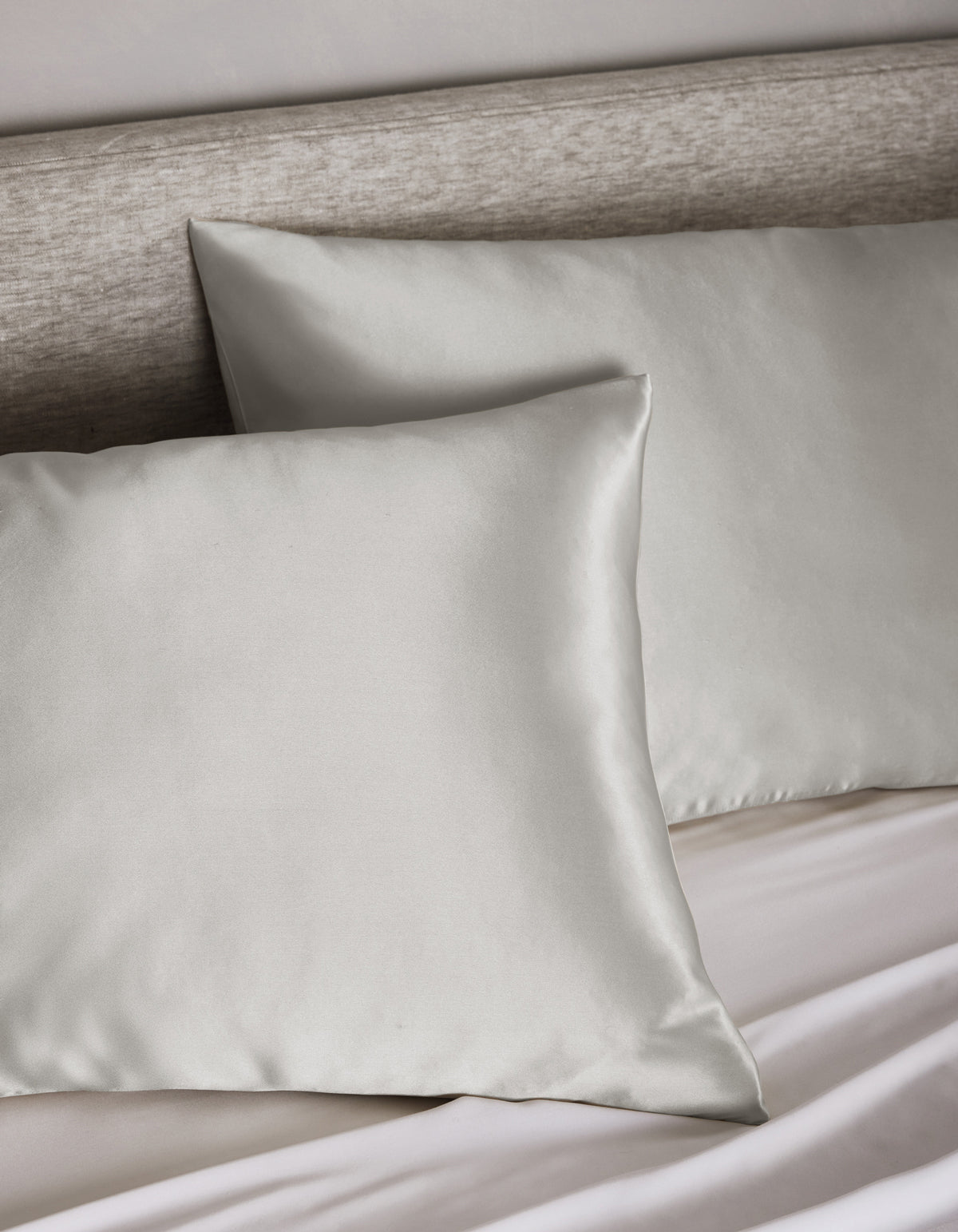 Two Cozy Earth Silk Pillowcases in light gray rest on a matching upholstered headboard and bed, with a neatly tucked white sheet visible below.