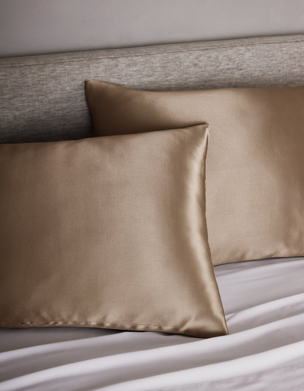 Two Cozy Earth Silk Pillowcases rest on a bed with a light gray upholstered headboard and white sheets.