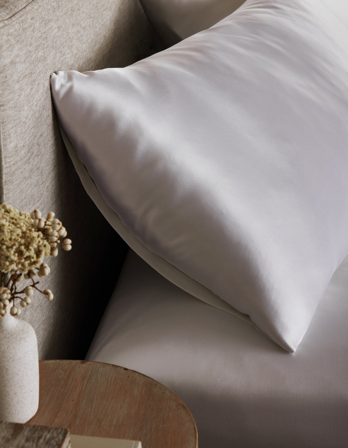 A Cozy Earth Silk Pillowcase sits on a bed with matching sheets by a beige headboard. In the foreground, a small wooden table holds a vase of dried flowers.