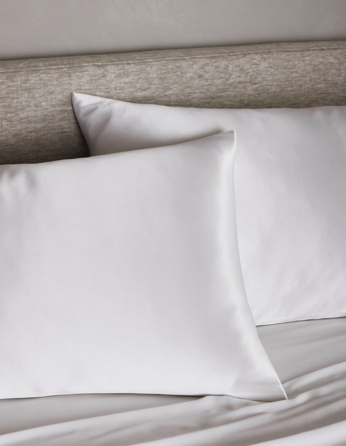 Two Cozy Earth Silk Pillowcases rest on a neatly made bed with a light gray upholstered headboard, creating a clean and minimalistic bedroom atmosphere.