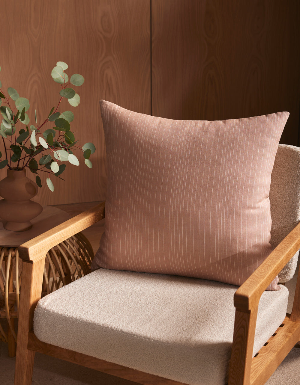A cozy wooden armchair with a textured beige cushion and a large Cozy Earth Striped Throw Pillow sits beside a small table topped with a brown vase of green eucalyptus. A wood-paneled wall forms the background.