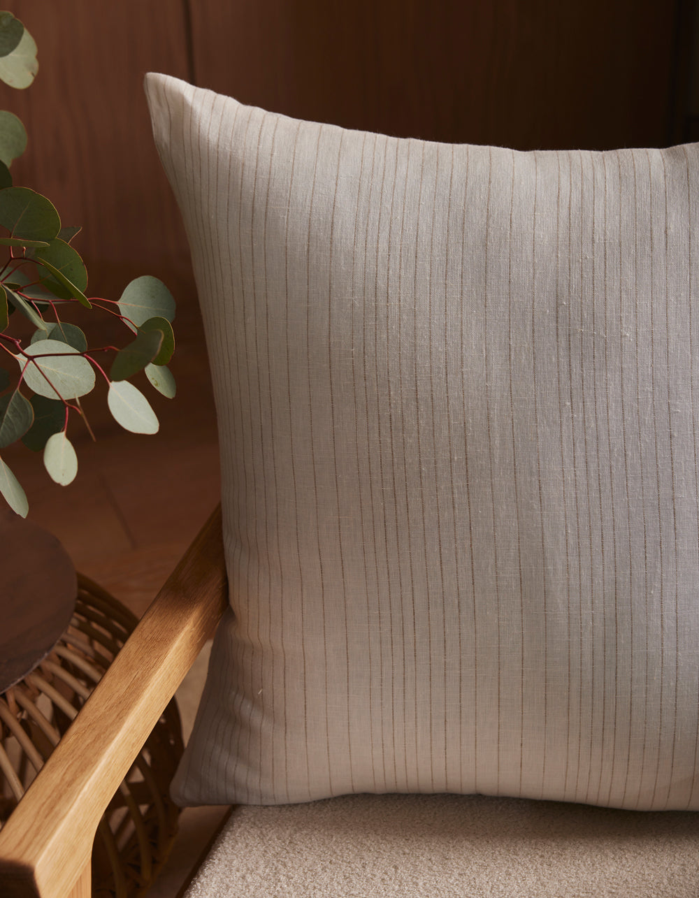 A close-up of the Cozy Earth Striped Throw Pillow on a light cushioned chair, next to a wooden armrest and a round table with eucalyptus leaves in a glass vase.