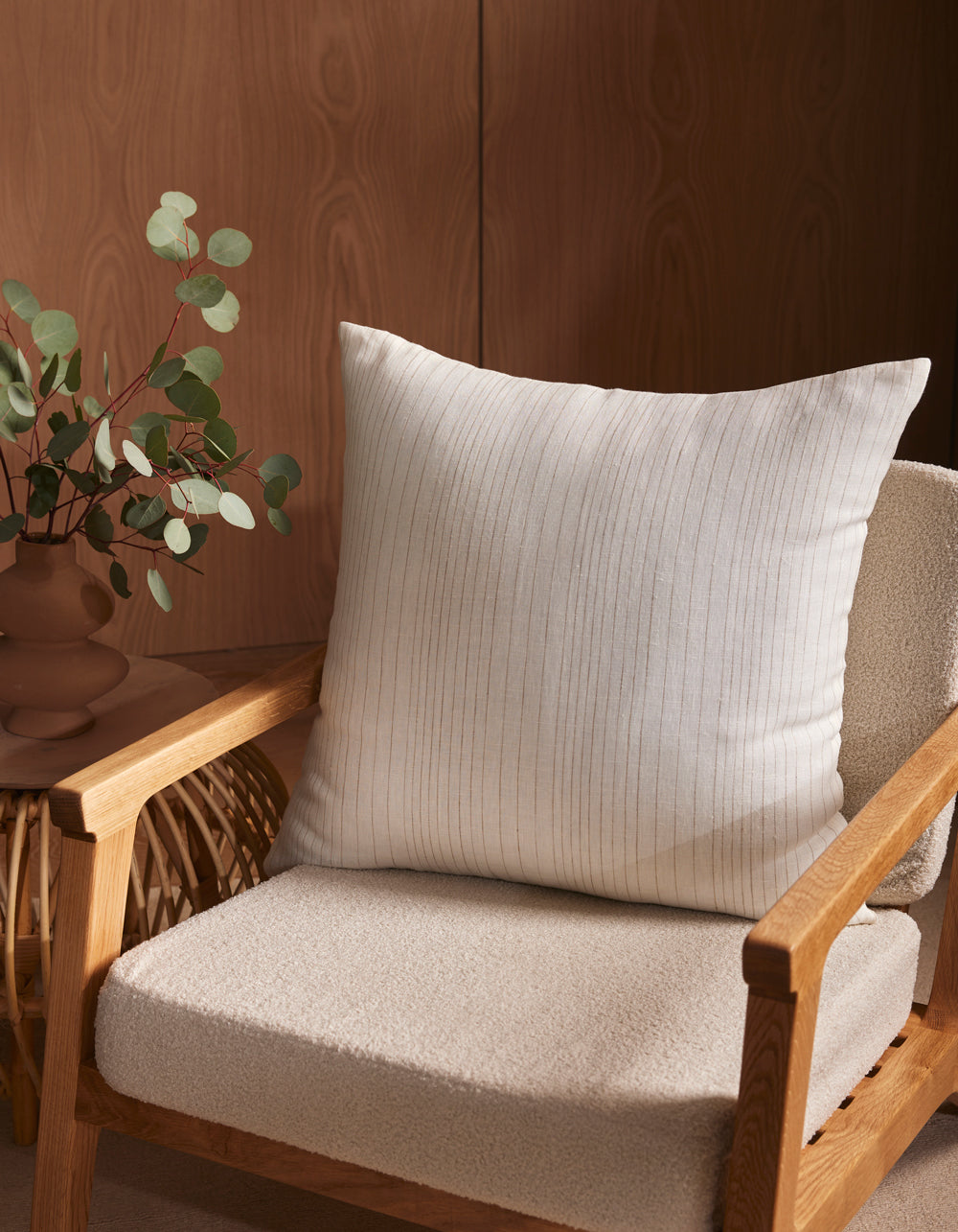 A Cozy Earth Striped Throw Pillow rests on a wooden armchair with a textured cream seat. Beside it, a small woven side table displays a brown vase filled with green eucalyptus branches.