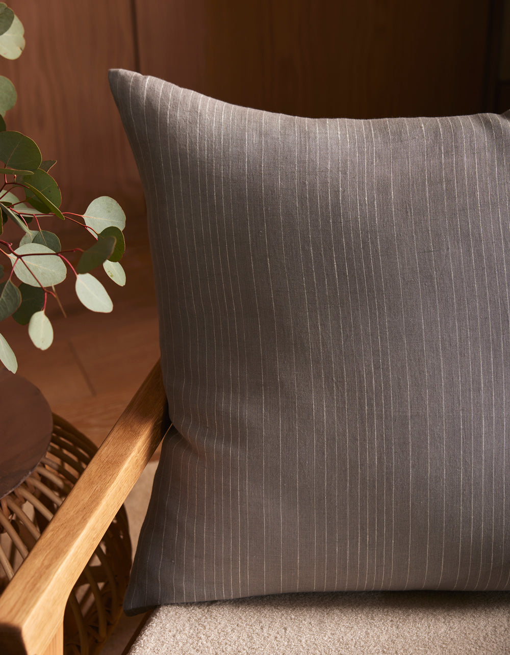 The Cozy Earth Striped Throw Pillow features gray fabric with thin white pinstripes, styled on a beige cushioned wooden chair beside a round woven table and green eucalyptus branches in a softly lit, warm-toned setting.