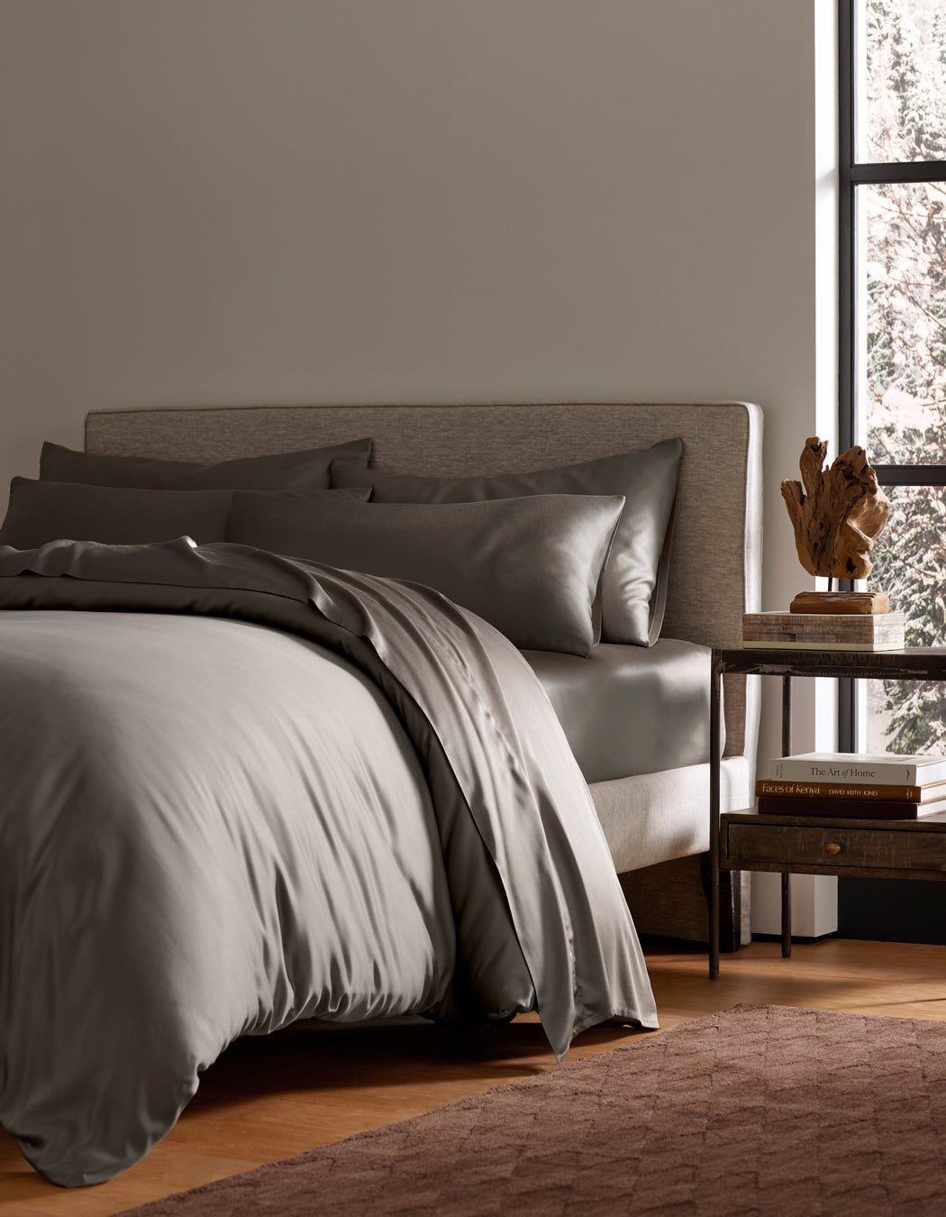 A modern bedroom with a neatly made bed featuring Cozy Earth's Bamboo Bedding Ultimate Bundle, two pillows, a beige upholstered headboard, a wooden nightstand with books and a sculpture, and a large window letting in natural light.|Color:Charcoal