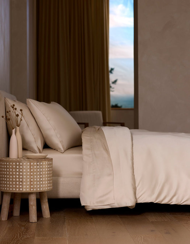 A neatly made bed with the Cozy Earth Bamboo Sheet Set in cream sits next to a round wooden nightstand holding two vases of dried flowers, as soft natural light streams through a window with beige curtains.