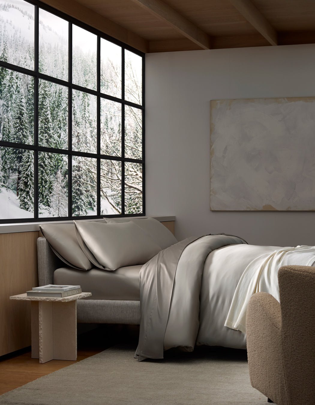 A cozy bedroom with a large window framing snowy pine trees outside. The bed features Cozy Earth's Bamboo Bedding Ultimate Bundle in gray, and there’s a beige chair and side table with books, creating a calm, minimalist retreat.