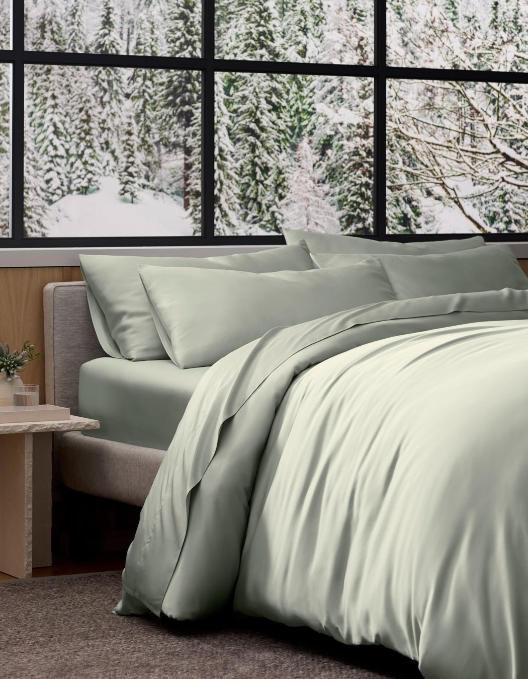 A neatly made bed with a light sage green Bamboo Duvet Cover by Cozy Earth sits by a window overlooking a snowy pine forest, with a small potted plant on the side table.
