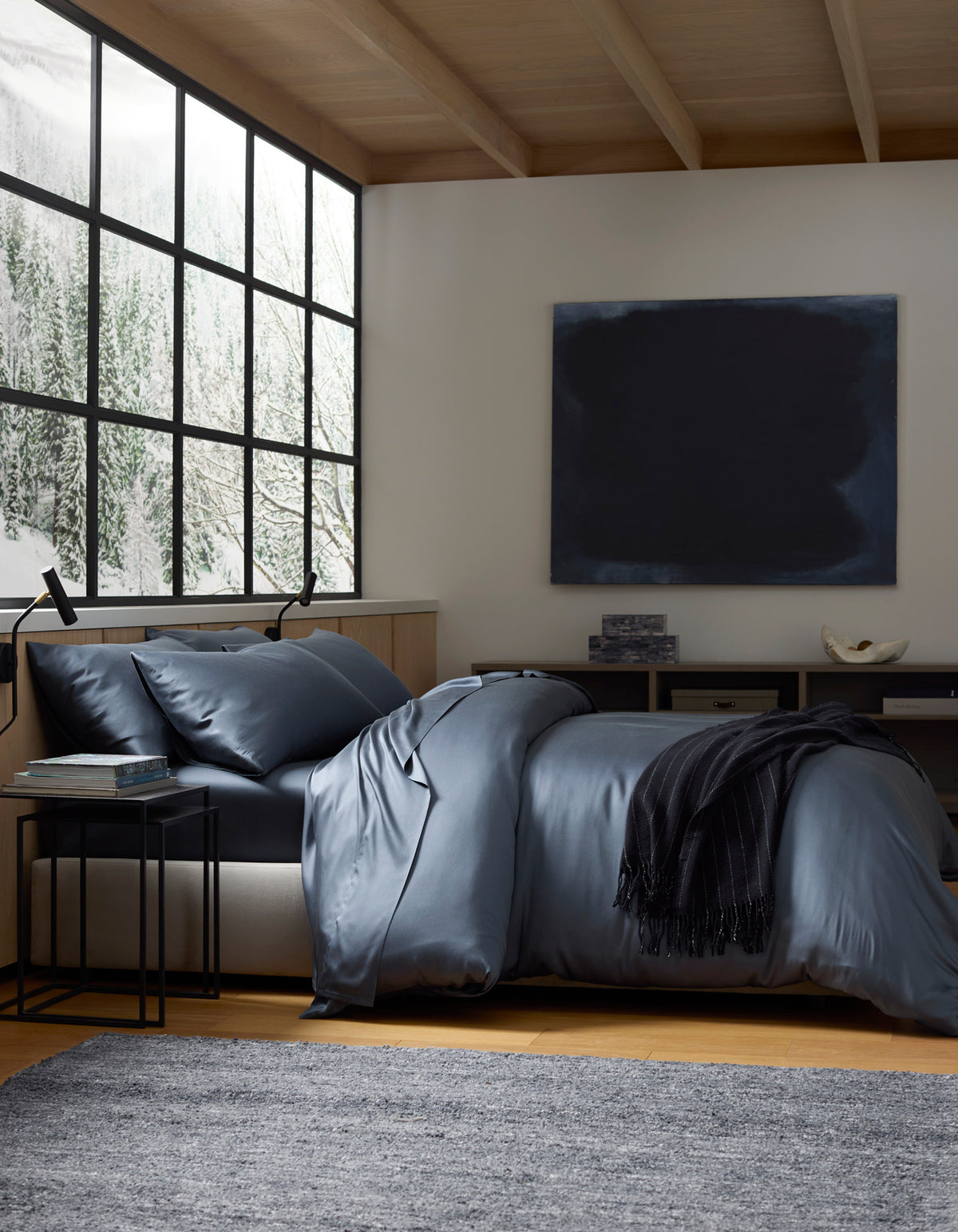 A modern bedroom with a large window showing snowy trees, a bed made with Cozy Earth's Bamboo Bedding Deluxe Bundle in dark blue, a black throw, side table, grey rug, and a dark abstract painting on the wall.