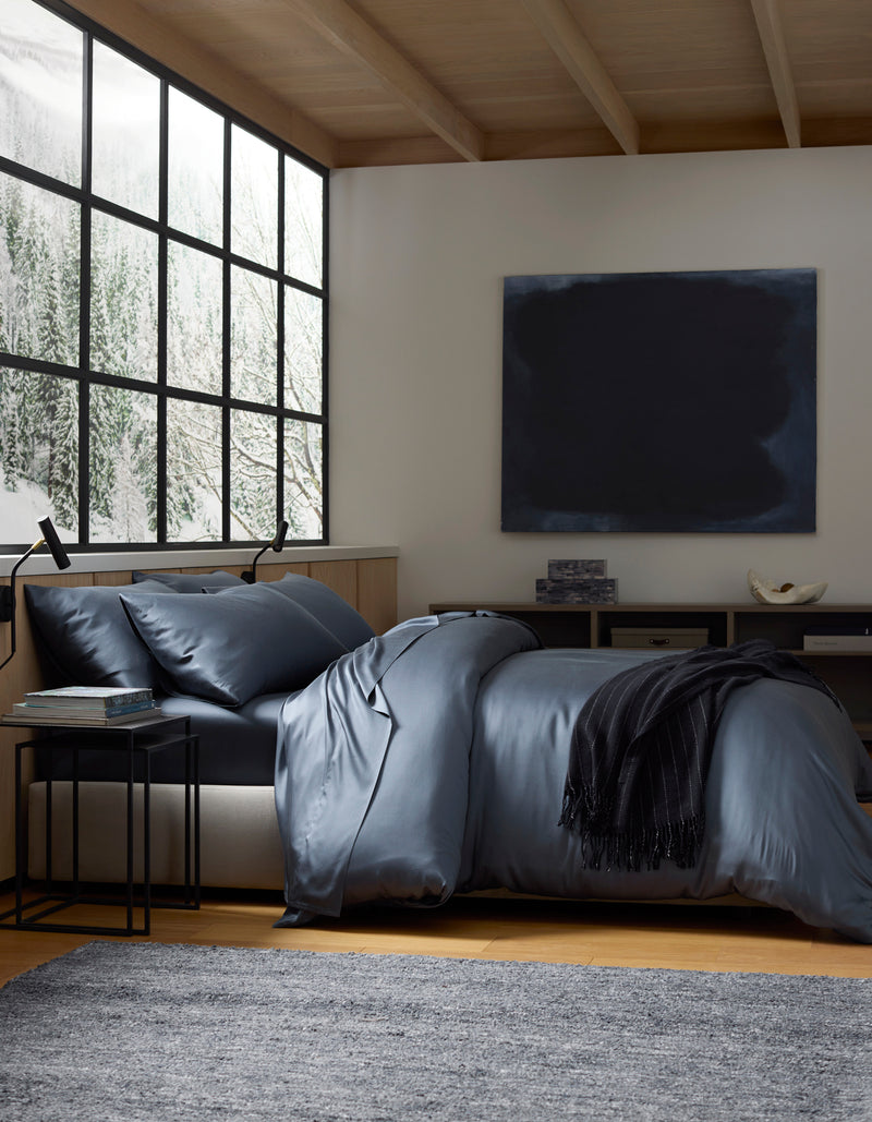 A modern bedroom with a large window showing snowy trees, a bed made with Cozy Earth's Bamboo Bedding Deluxe Bundle in dark blue, a black throw, side table, grey rug, and a dark abstract painting on the wall.