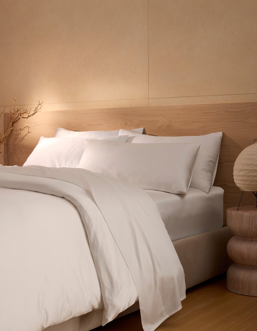 A minimalist bedroom featuring the Cozy Earth Bamboo Bedding Ultimate Bundle—white sheets, pillows, and comforter—next to a wooden nightstand with a round lamp and decorative branch in soft lighting.