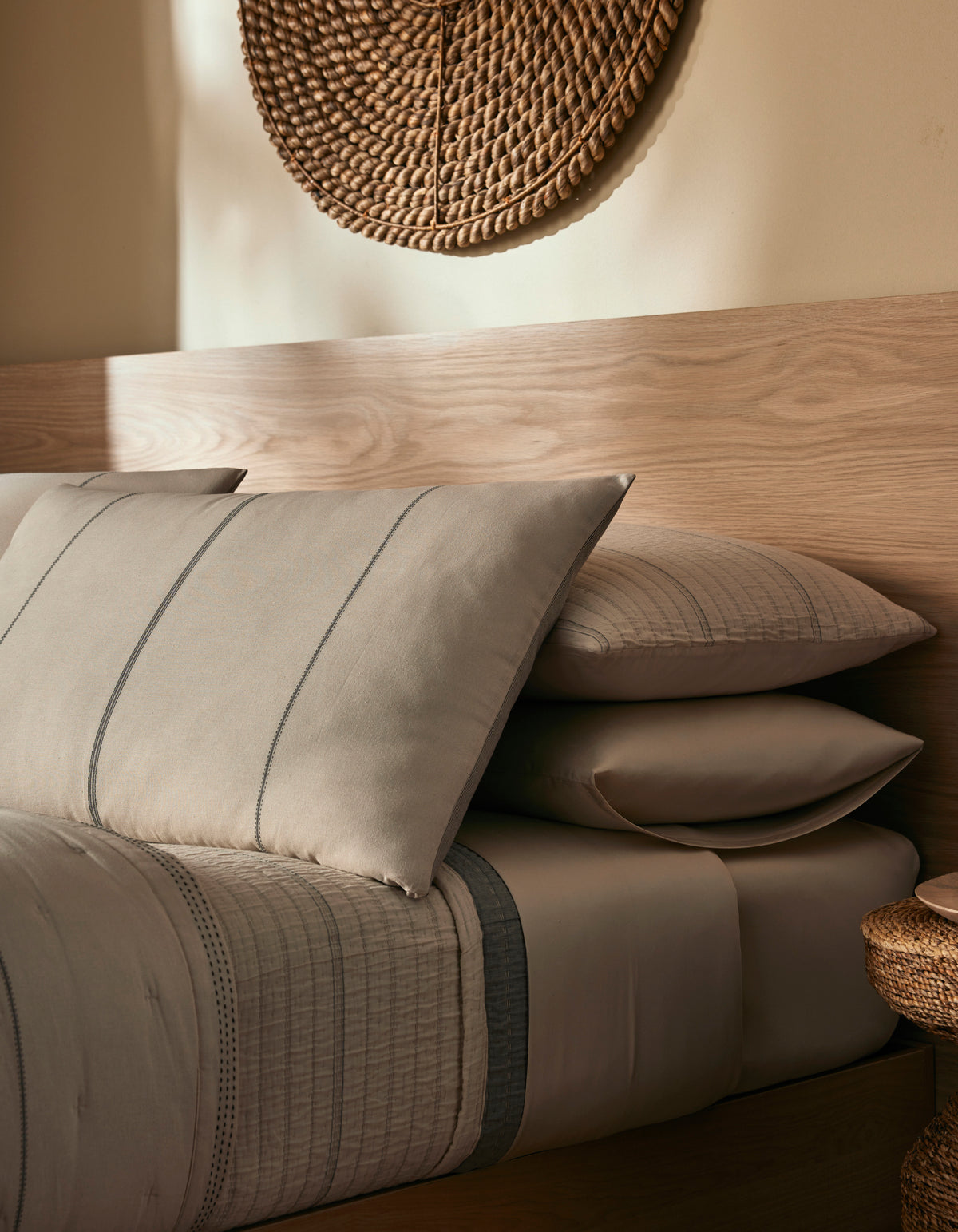A cozy bedroom featuring the Cozy Earth Baja Double Sided Sham with taupe and gray bedding, stacked pillows, a light wood headboard, and a woven wall piece above, all softly lit for a warm ambiance.