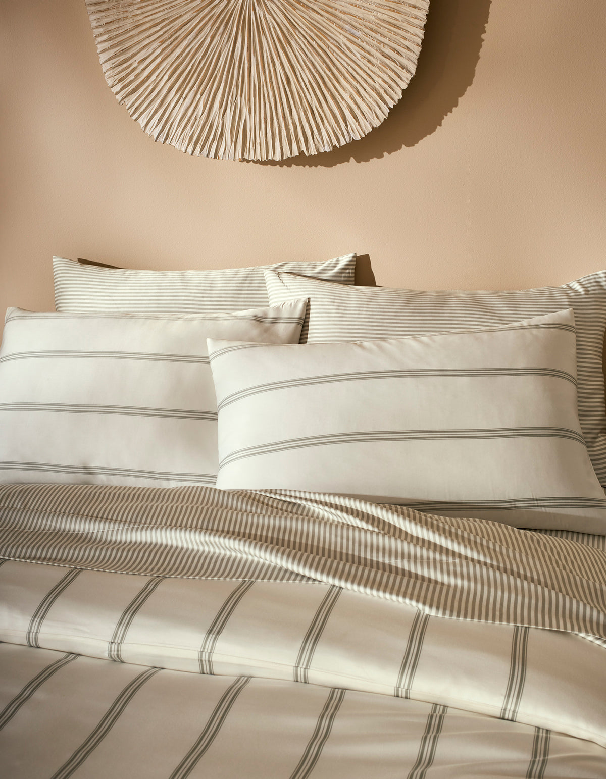 A neatly made bed features Cozy Earth Bamboo Pillowcases in cream and light gray stripes, accented by matching bedding and set against a beige wall with a textured, fan-shaped decoration above the headboard.