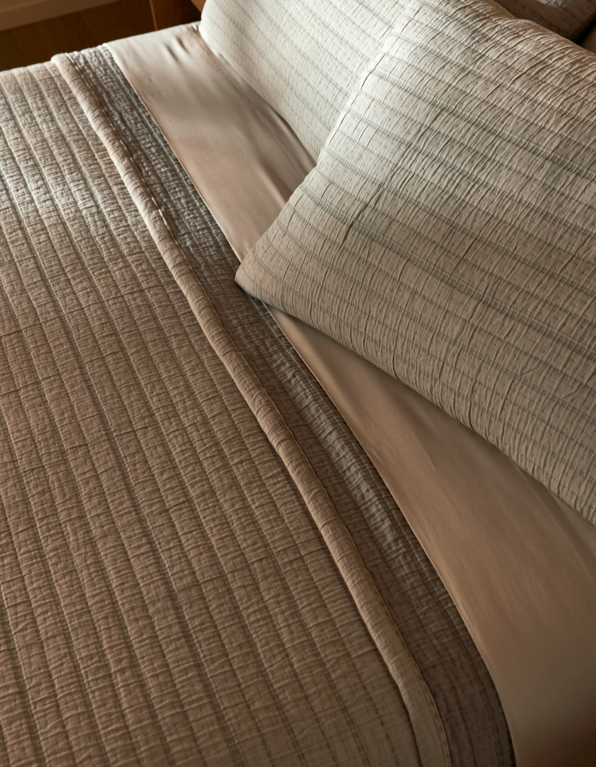 Close-up of a neatly made bed featuring the Cozy Earth Baja Matelasse Coverlet in light gray with matching pillows, accented by a smooth white sheet underneath. The bedding looks clean and inviting.