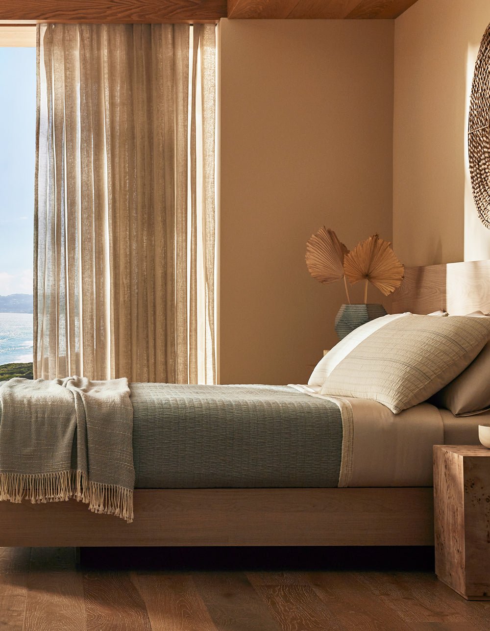 A sunlit, minimalist bedroom featuring the Cozy Earth Baja Matelasse Coverlet on a wooden bed, with neutral decor, a woven wall hanging, nightstand, dried leaves, and sheer curtains framing a sea view.