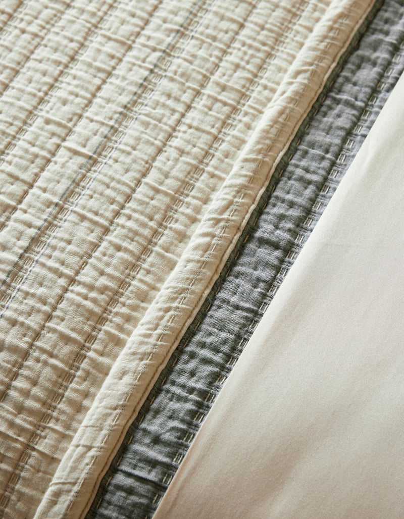 A close-up of layered bedding featuring the Cozy Earth Baja Matelasse Coverlet in beige, a gray quilted blanket, and a smooth cream-colored sheet.