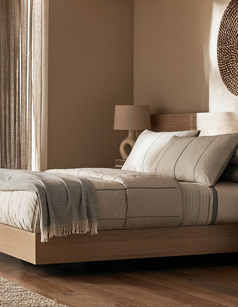 Minimalist bedroom featuring a light wood platform bed, beige bedding, the Cozy Earth Baja Tassel Throw in gray, round woven wall decor, a modern beige lamp, and soft natural light through sheer curtains.