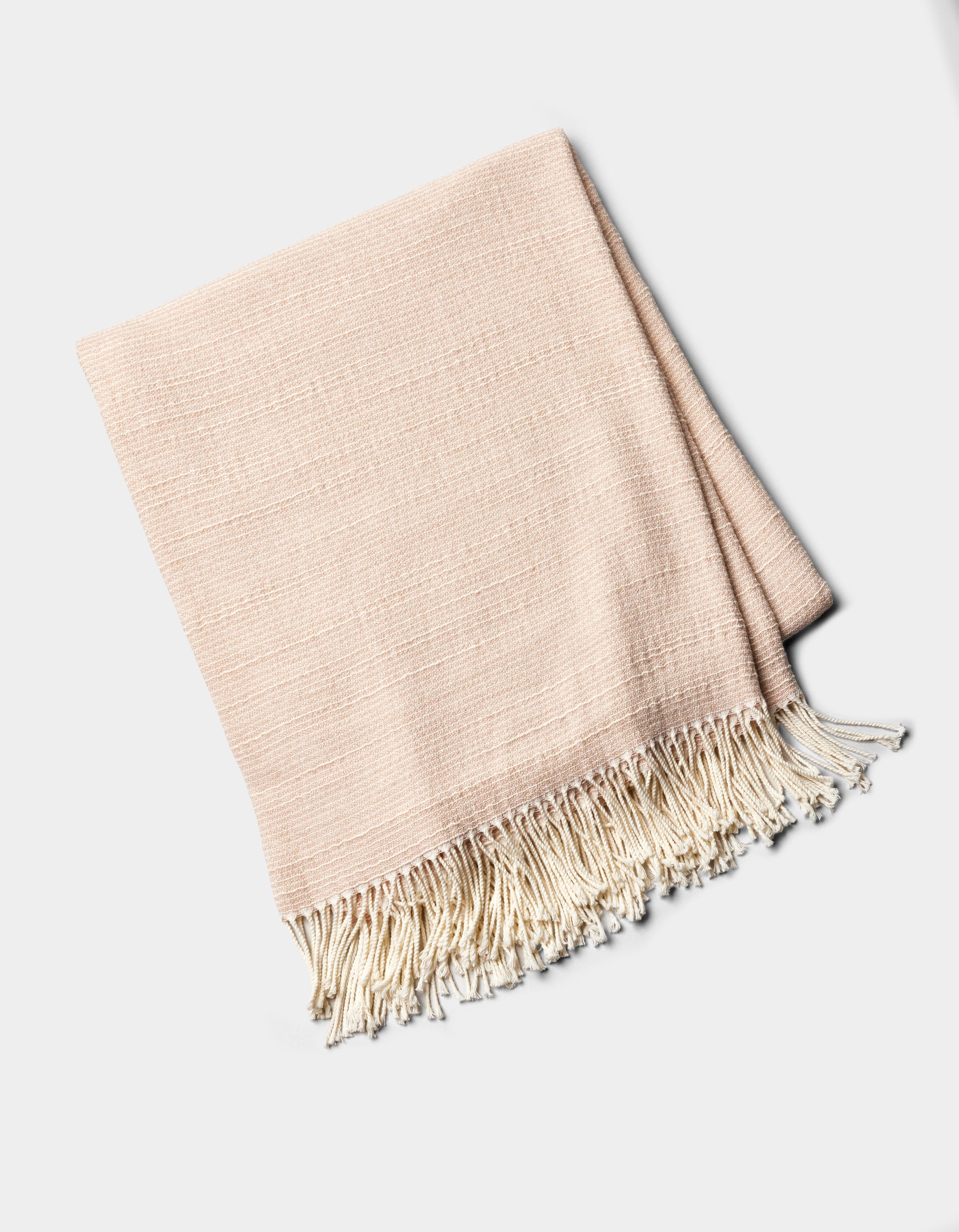 The Cozy Earth Baja Tassel Throw is a folded, light beige woven blanket with cream fringe on one end, shown on a plain white background.|Color:Himalayan Salt