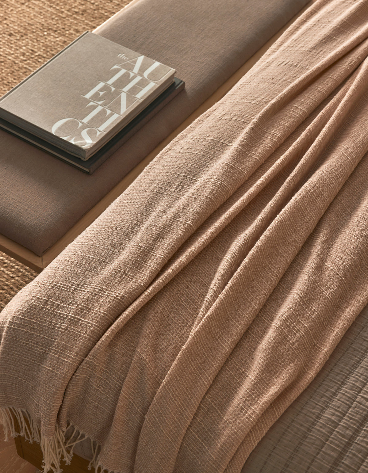 The Baja Tassel Throw by Cozy Earth, in beige with a textured finish, is draped over a neutral-toned bench beside two stacked books (one titled "AUTHENTICS"), creating a cozy, minimalistic look.