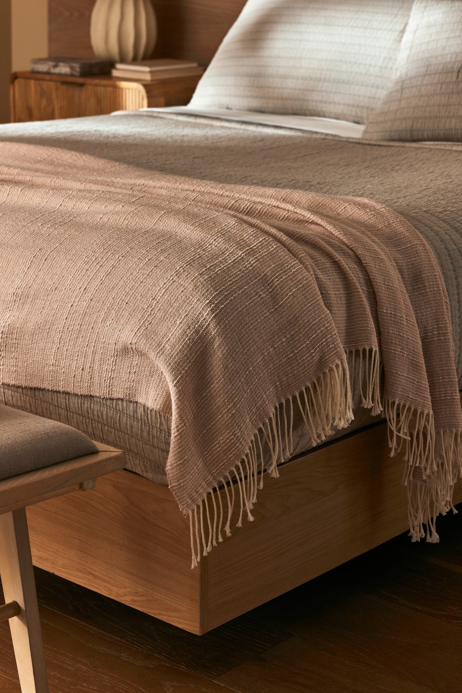 A close-up of a neatly made bed with the Cozy Earth Baja Tassel Throw draped at the foot. The wooden frame and neutral bedding create a cozy, minimalist vibe, complemented by the throw’s light texture.|Color:Himalayan Salt