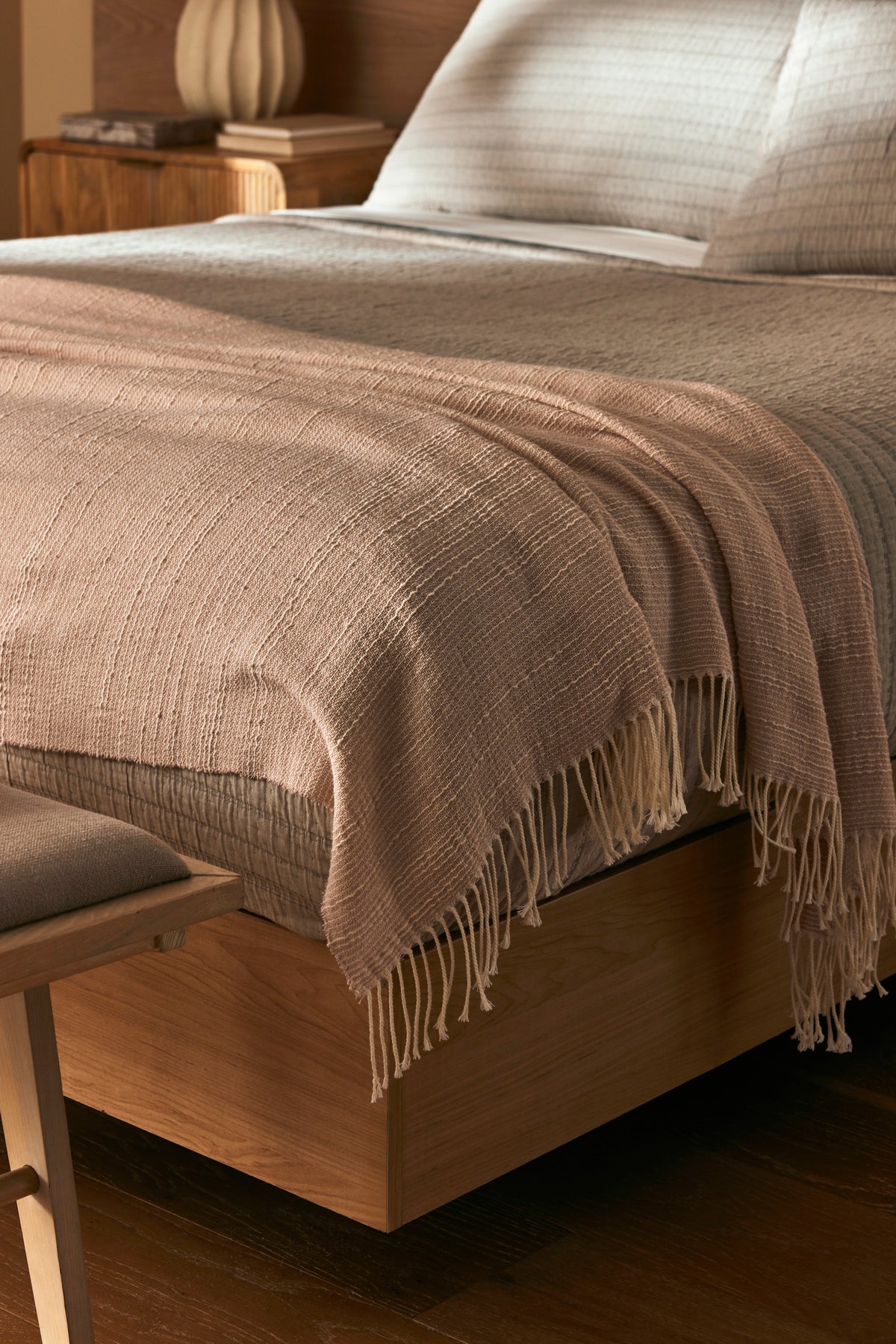A close-up of a neatly made bed with the Cozy Earth Baja Tassel Throw draped at the foot. The wooden frame and neutral bedding create a cozy, minimalist vibe, complemented by the throw’s light texture.
