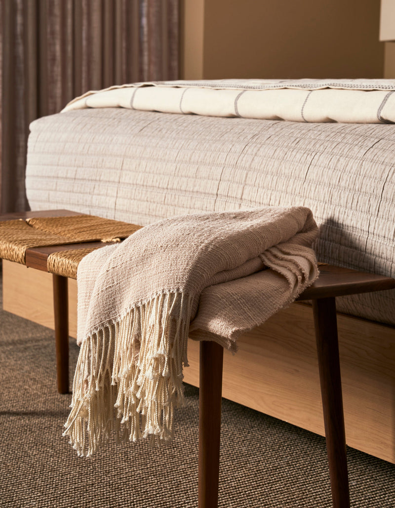The Cozy Earth Baja Tassel Throw, a beige woven blanket with fringe, is draped over a wooden bench at the foot of a bed with a textured light bedspread, complementing the room's warm neutral tones and soft lighting.