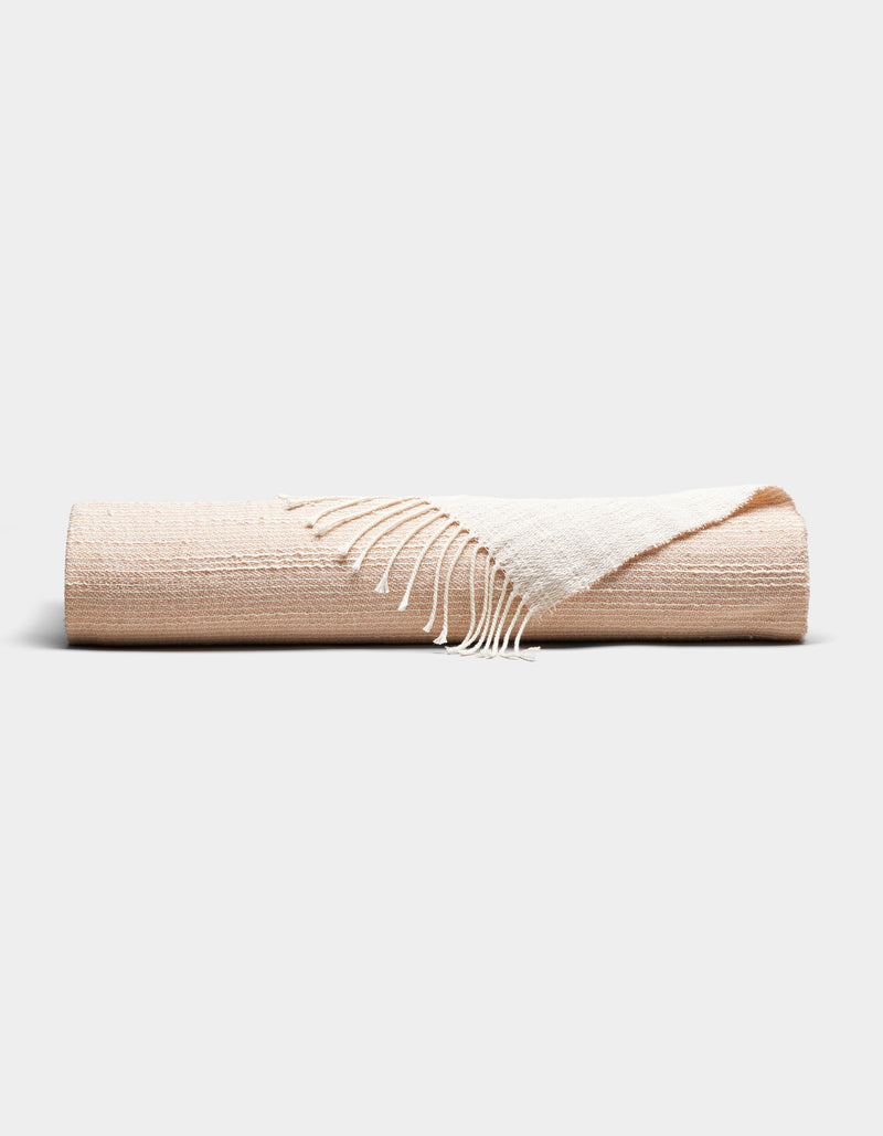 The Cozy Earth Baja Tassel Throw is a neatly rolled beige blanket with white fringed edges, showcased on a plain light gray background.