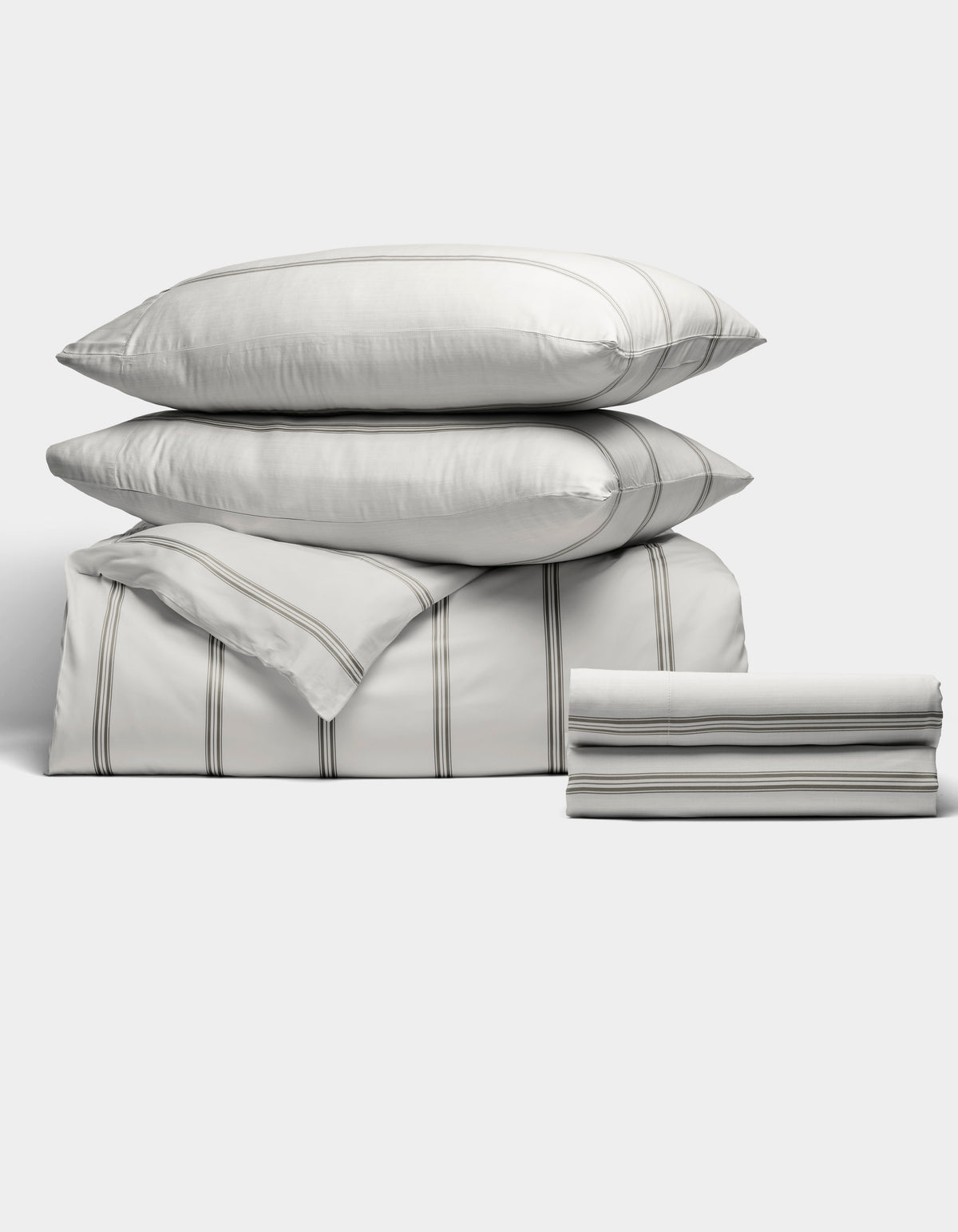 The Bamboo Bedding Deluxe Bundle by Cozy Earth features two pillows, a folded comforter, and a folded sheet in white with thin gray stripes, all neatly stacked against a plain light background.