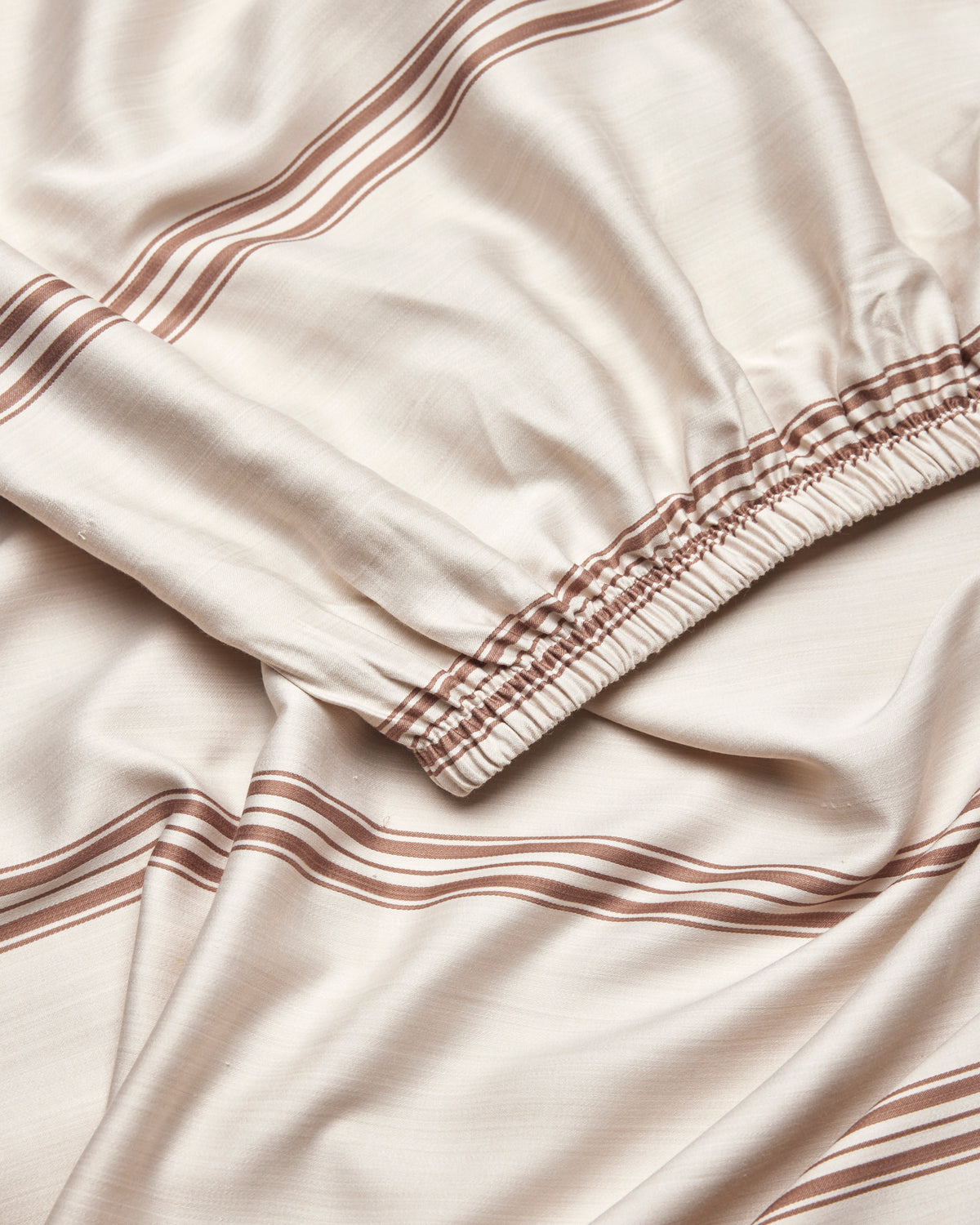 A close-up of Cozy Earth's Bamboo Fitted Sheet in beige, featuring brown horizontal stripes and an elastic gathered edge, displayed in soft folds.