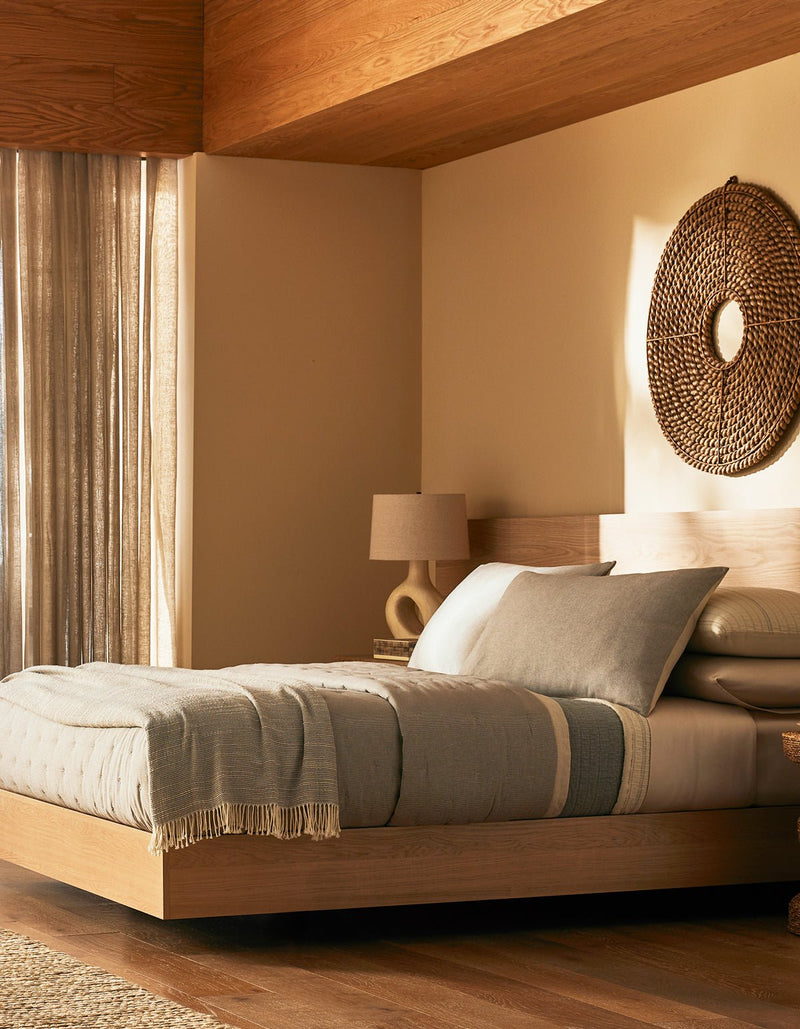 A modern bedroom features Cozy Earth's Baja Quilt Bundle on a wooden platform bed, with neutral tones, a woven wall hanging, a beige lamp on a nightstand, and sunlight filtering through sheer curtains onto wood floors.