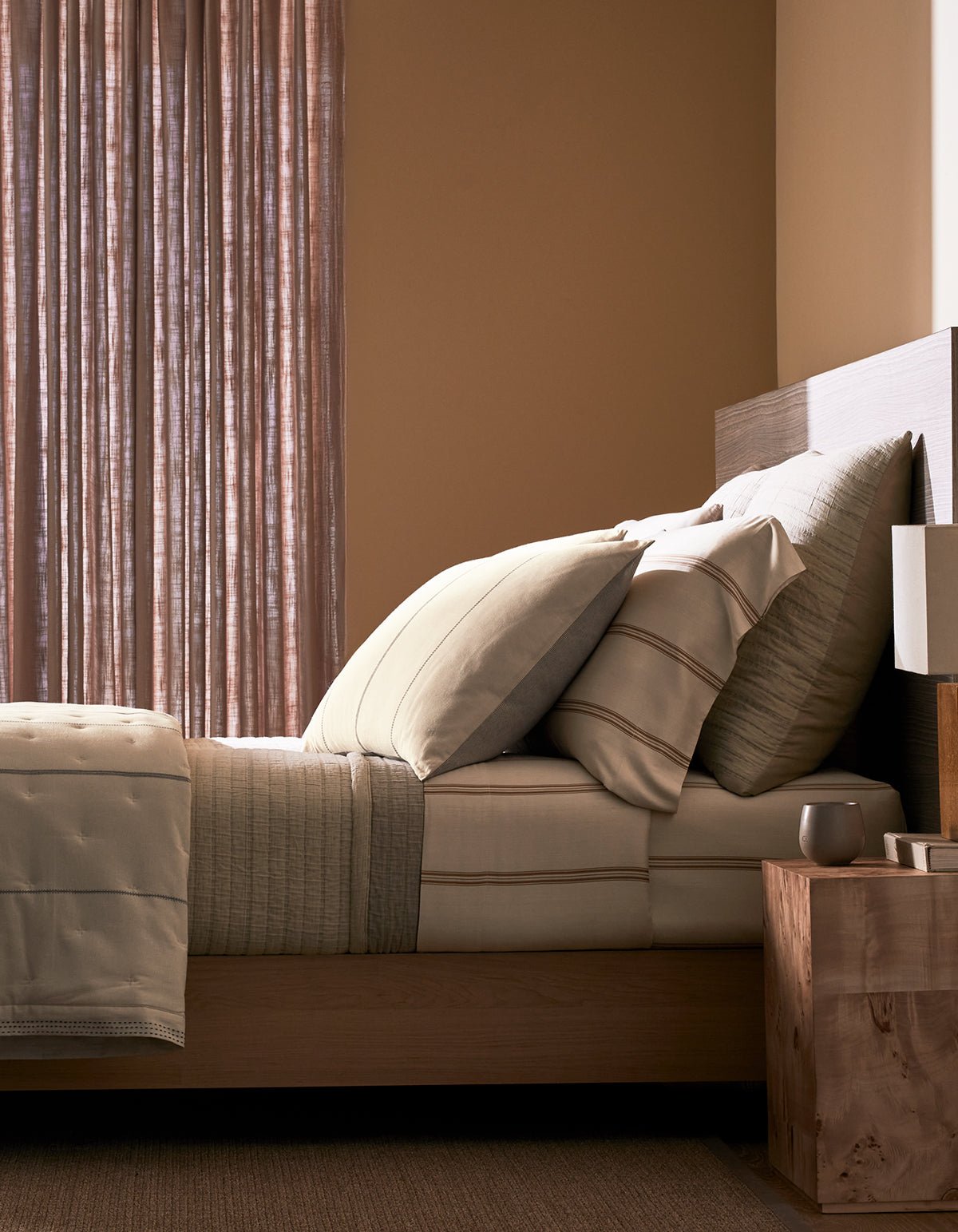 A neatly made bed featuring Cozy Earth's Baja Matelasse Bundle sits beside a wooden nightstand with a lamp and cup, set in a softly lit room with sheer curtains and beige walls.