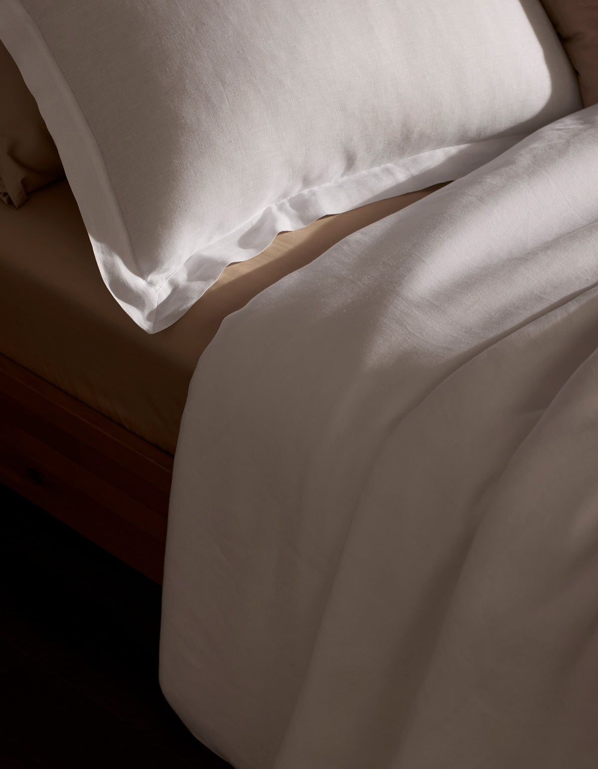 A neatly made bed features the Cozy Earth Linen Duvet Set—a white pillow and sheet atop a beige fitted sheet—all bathed in soft natural light.