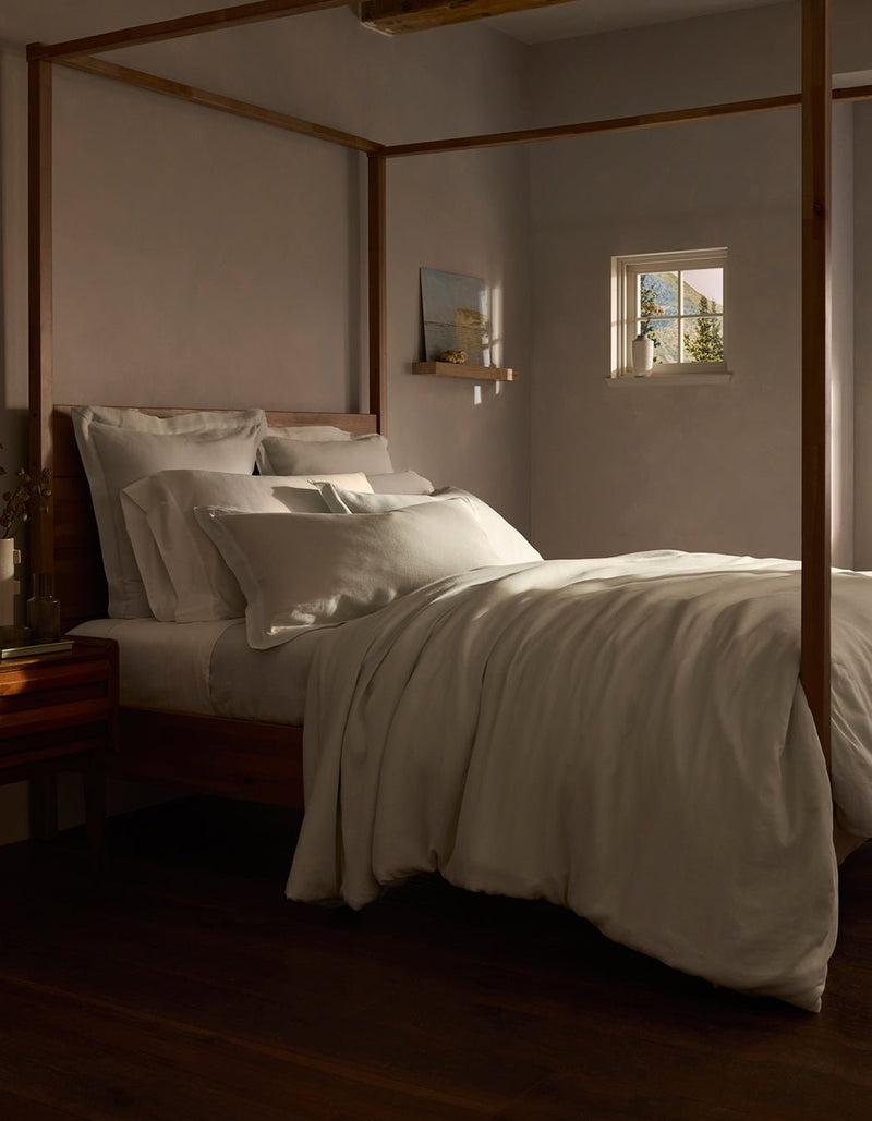 A cozy bedroom features a wooden four-poster bed with layered white bedding, including the Linen Duvet Cover by Cozy Earth. Warm light from a small window softly illuminates the neutral tones, creating an inviting atmosphere.