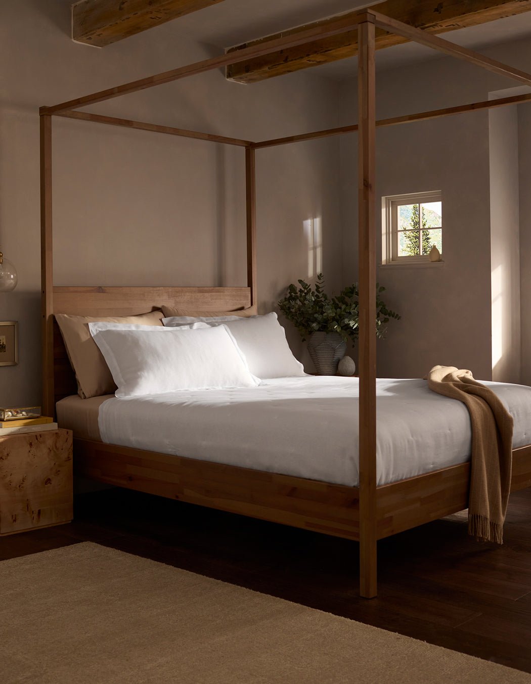 A cozy bedroom features a wooden four-poster bed with white bedding and a Cozy Earth Linen Quilt draped at the foot. Soft natural light fills the space, and a vase with greenery rests on the bedside table.