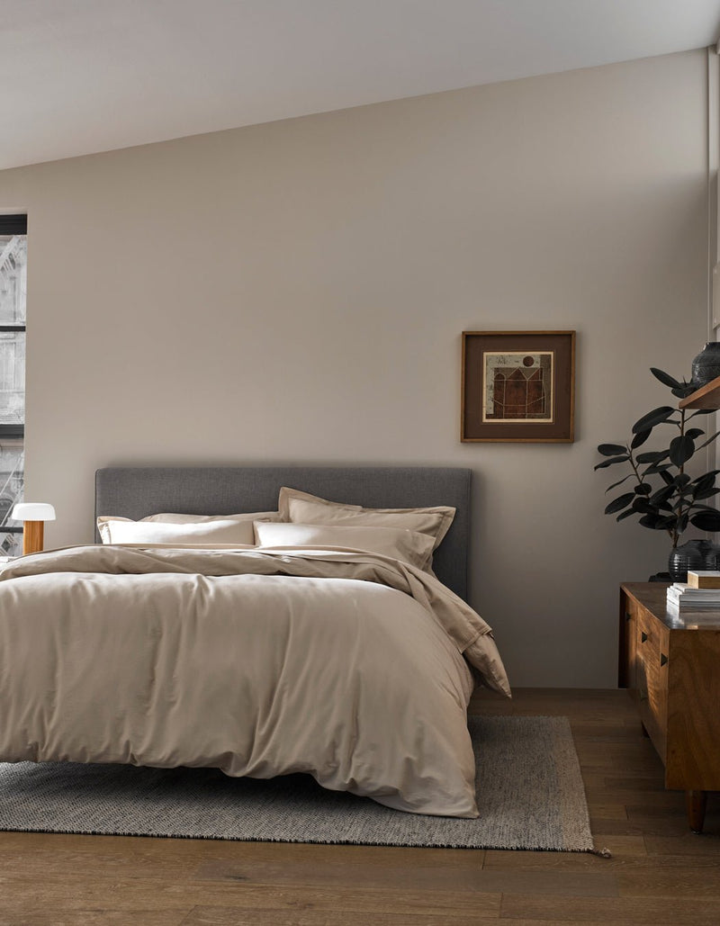 A modern bedroom features a gray upholstered bed with Cozy Earth's Soft-Wash Cotton Duvet Cover in beige, a wooden nightstand holding a plant and lamp, a framed picture on a light-colored wall, all atop wood flooring with a neutral rug.