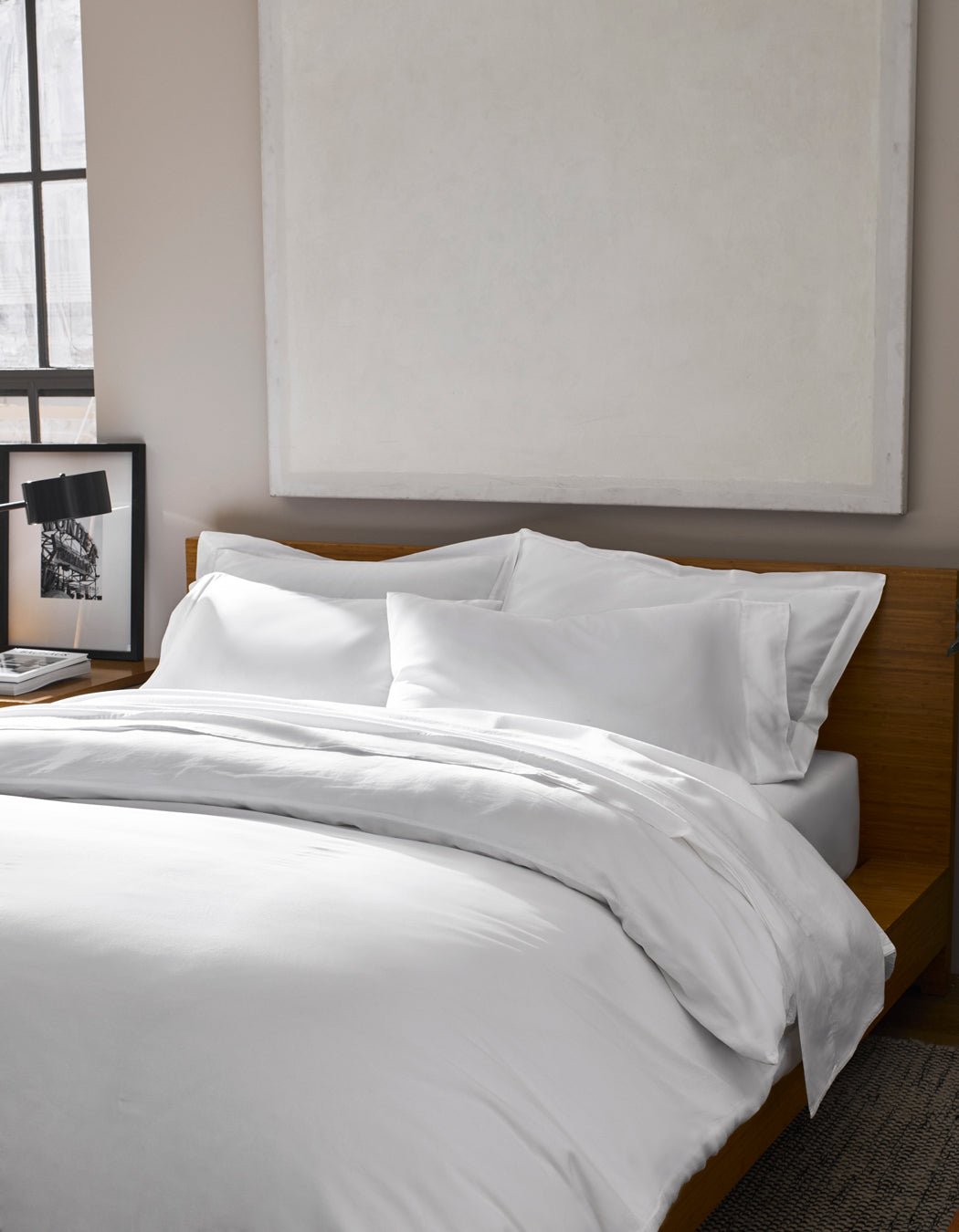 A Soft-Wash Cotton Duvet Cover by Cozy Earth dresses the bed with white sheets against a wooden headboard. Above, a large abstract painting hangs, while a framed photo sits on the bedside table near the window.