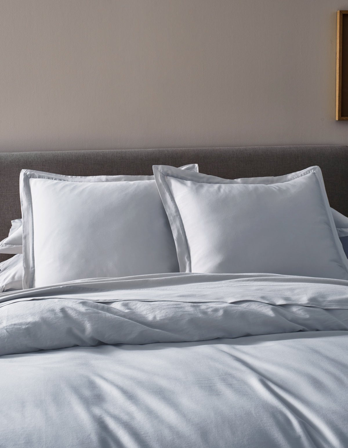 A neatly made bed features Cozy Earth’s Soft-Wash Cotton Duvet Cover, a gray upholstered headboard, two large white pillows, and white sheets in a softly lit bedroom.