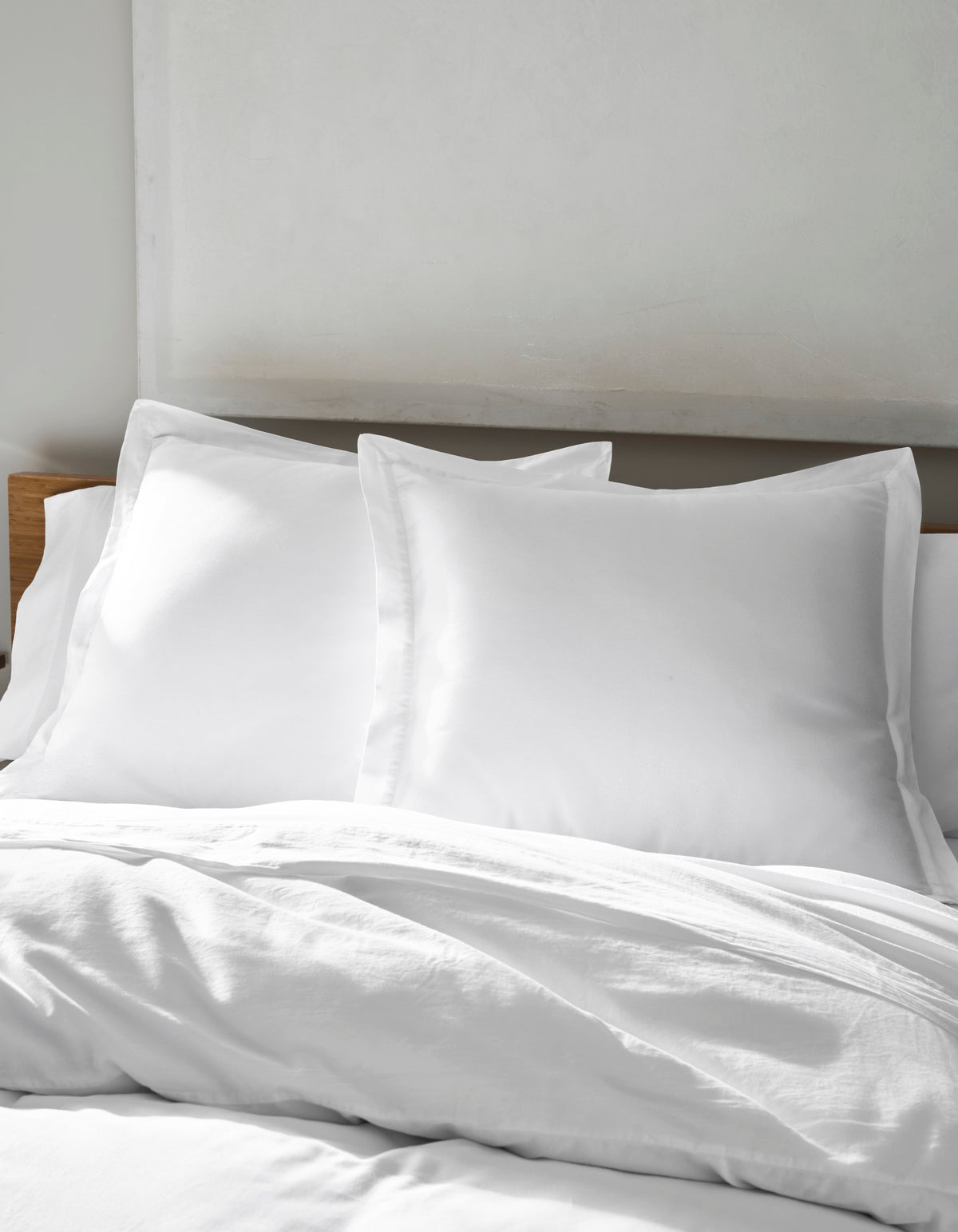 A neatly made bed features Cozy Earth's Soft-Wash Cotton Shams—large, fluffy white pillows with a white duvet—set against a light-colored headboard and wall.