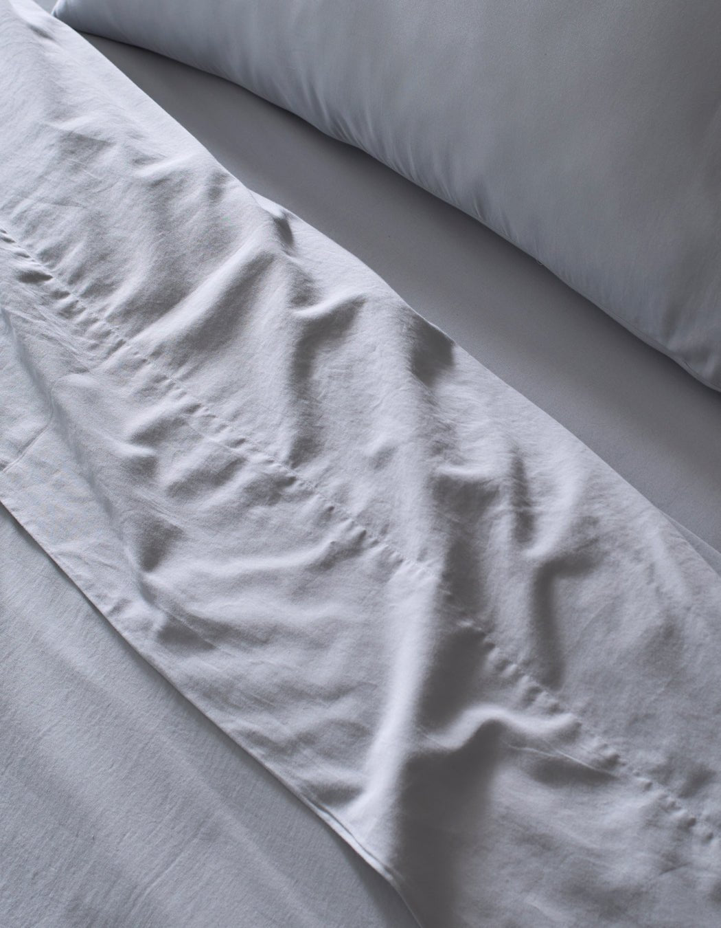 Close-up of a neatly made bed featuring Cozy Earth's Soft-Wash Cotton Sheet Set in light grey. Slight wrinkles add texture to the soft, clean fabric, giving the bedding an inviting look.