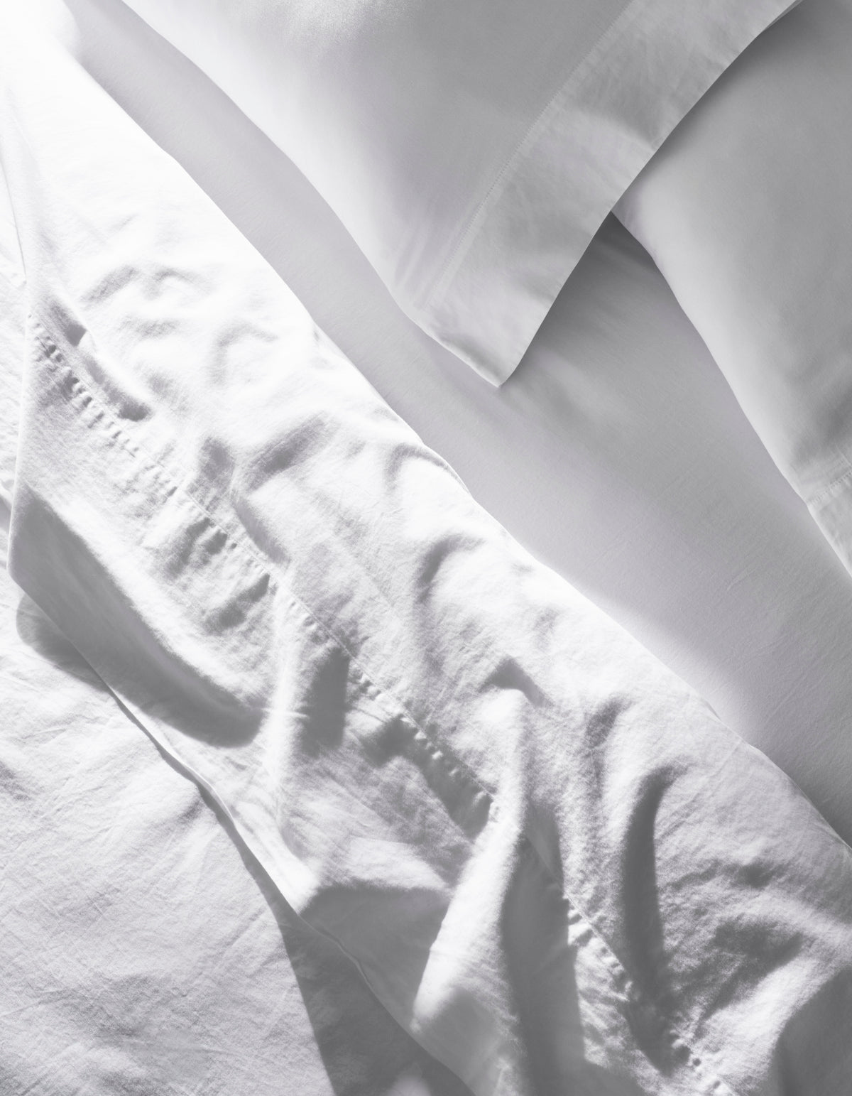 A close-up of the Cozy Earth Soft-Wash Cotton Bedding Core Bundle shows slightly wrinkled white sheets and pillowcases, with soft natural light casting gentle shadows and highlights on the fabric.