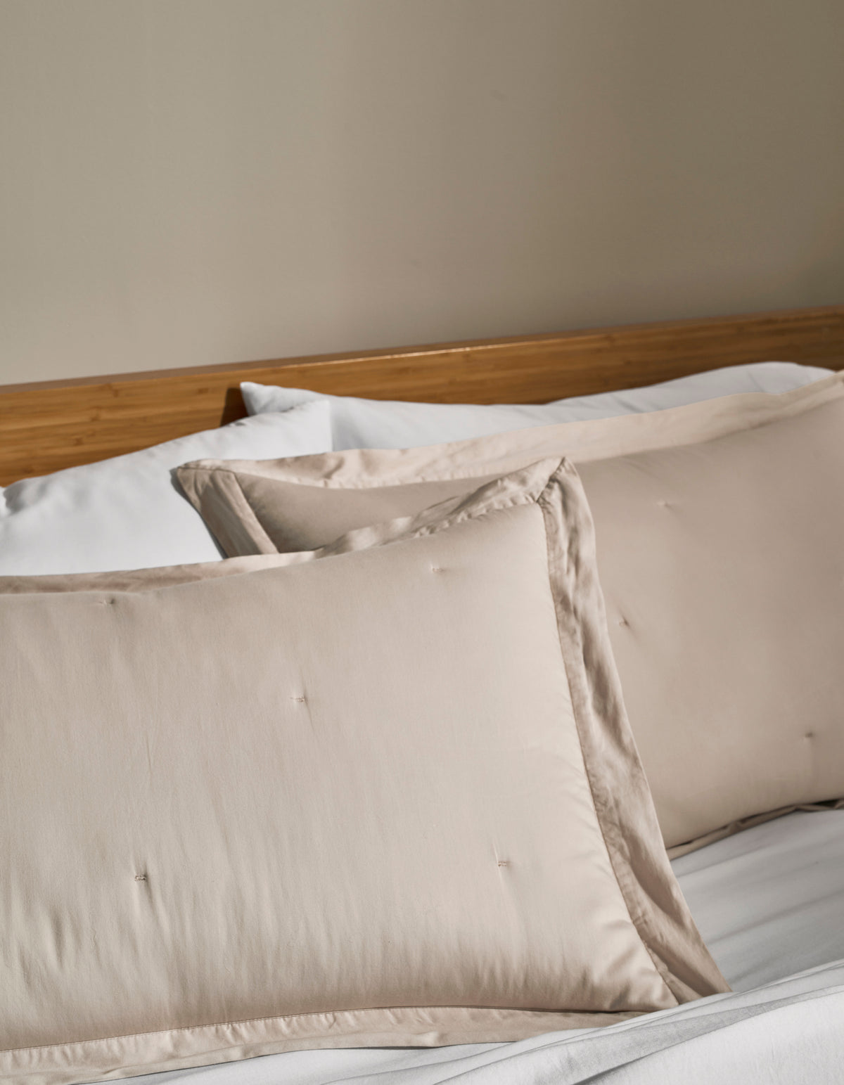 Two Cozy Earth Soft-Wash Cotton Quilted Shams in cream rest on a neatly made bed with white sheets and a wooden headboard against a beige wall.