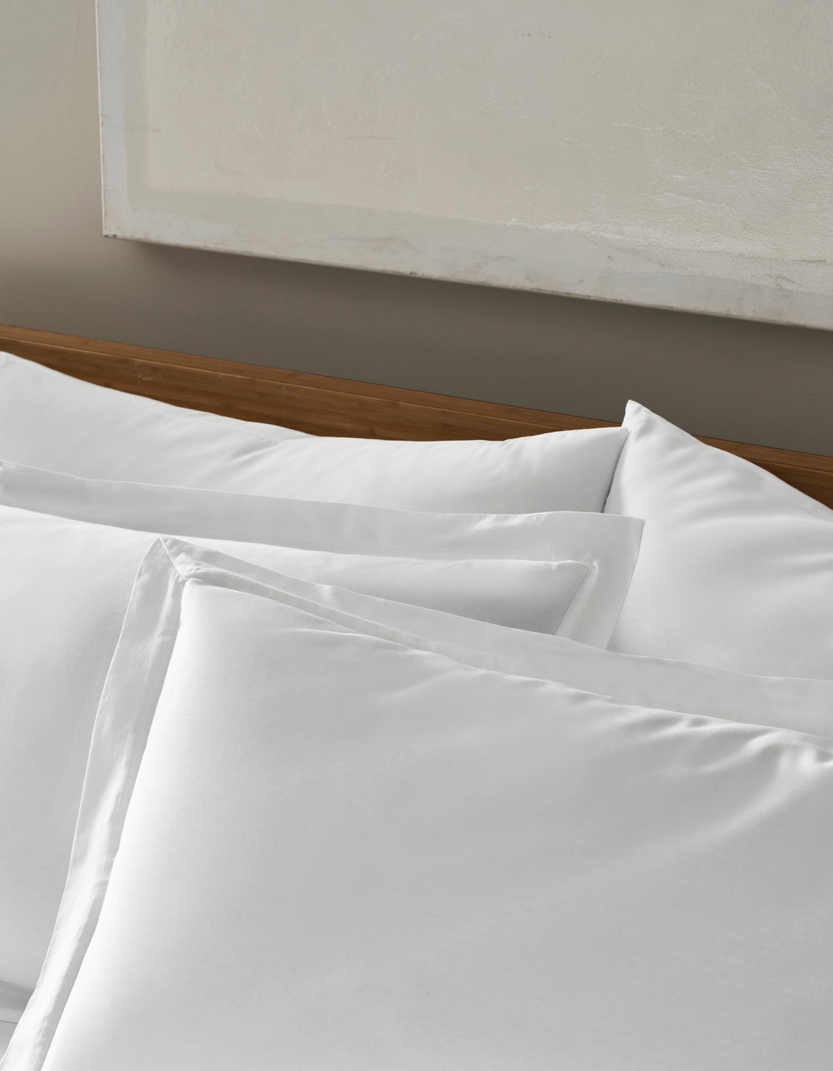 Four Soft-Wash Cotton Shams by Cozy Earth with matching pillowcases are neatly arranged on a bed with a wooden headboard, set against a neutral-toned wall accented by abstract artwork.