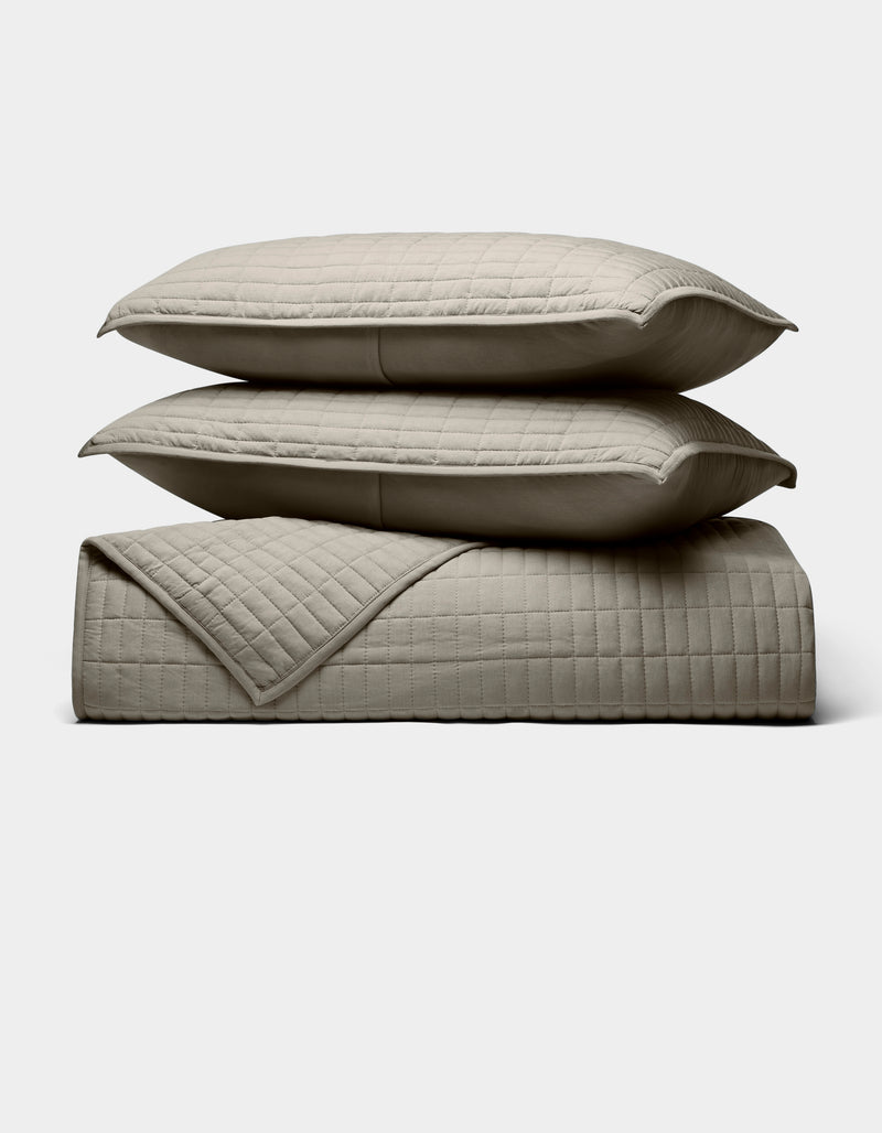 Two beige Cozy Earth Bamboo Jersey Quilt Bundle pillows are stacked atop a matching quilted blanket, all neatly arranged against a plain light background.