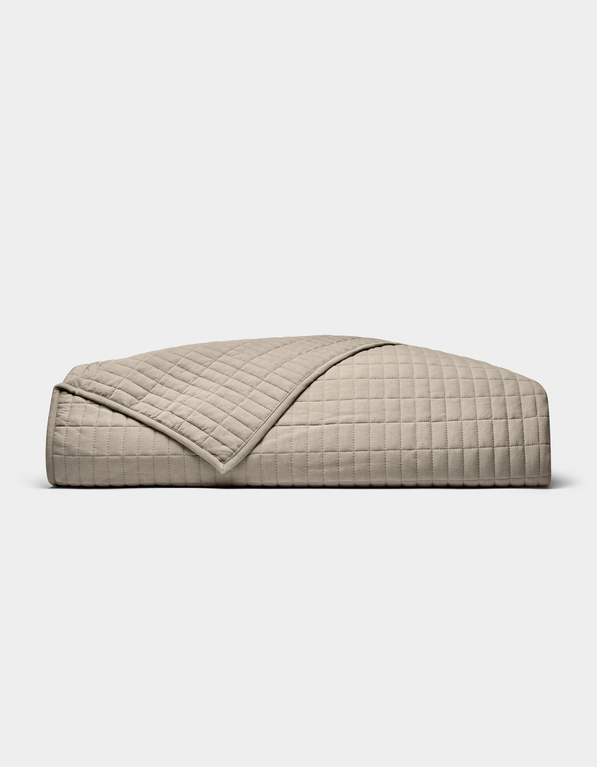 The Cozy Earth Bamboo Jersey Quilt, featuring a beige, quilted grid pattern, is neatly folded on a white background. A turned-down corner highlights its soft, luxurious texture.|Color:Driftwood