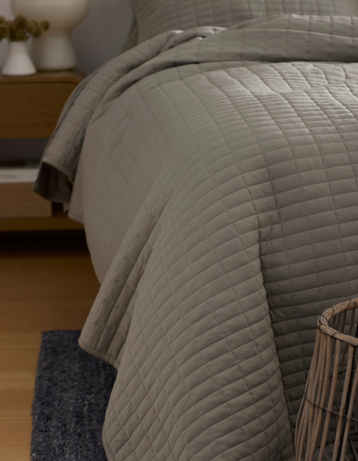 A close-up of a neatly made bed features the Cozy Earth Bamboo Jersey Quilt in light gray, draped over the side. Also visible are part of a wicker basket, a dark rug, and a wooden nightstand in this cozy bedroom corner.