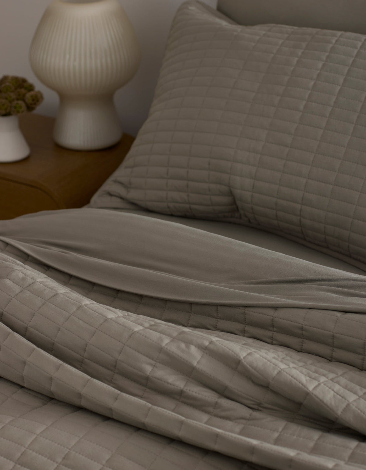 A neatly made bed features the Bamboo Jersey Quilt by Cozy Earth in taupe with matching pillows, next to a wooden nightstand topped with a white ribbed lamp and a small flower vase.