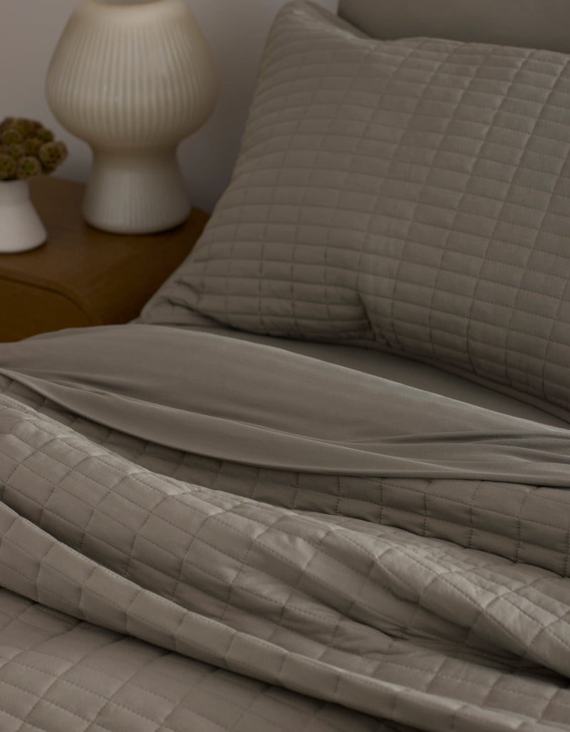 A neatly made bed features the Bamboo Jersey Quilt by Cozy Earth in taupe with matching pillows, next to a wooden nightstand topped with a white ribbed lamp and a small flower vase.