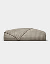 The Cozy Earth Bamboo Jersey Quilt is shown folded against a white background, with one corner turned down to display its beige color, grid pattern, and detailed stitching.|Color:Stone