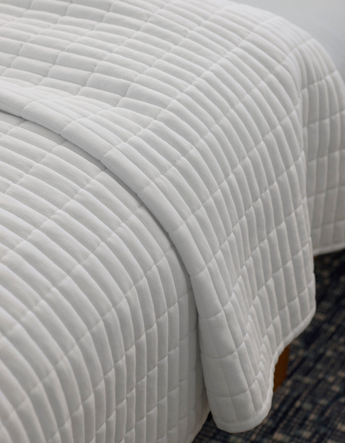 A close-up of a neatly made bed with the Cozy Earth Bamboo Jersey Quilt, featuring a white, square-patterned design, draped over the edge atop a dark patterned rug.