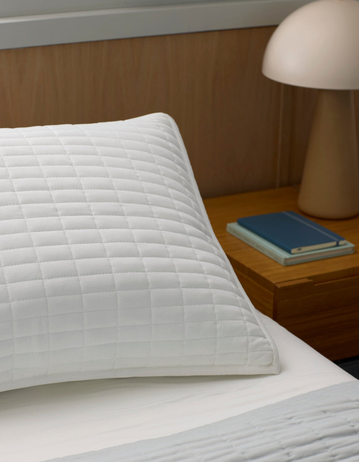 A close-up of a white Bamboo Jersey Quilted Sham by Cozy Earth, resting on a bed beside a wooden nightstand with a beige lamp and two neatly stacked closed books.